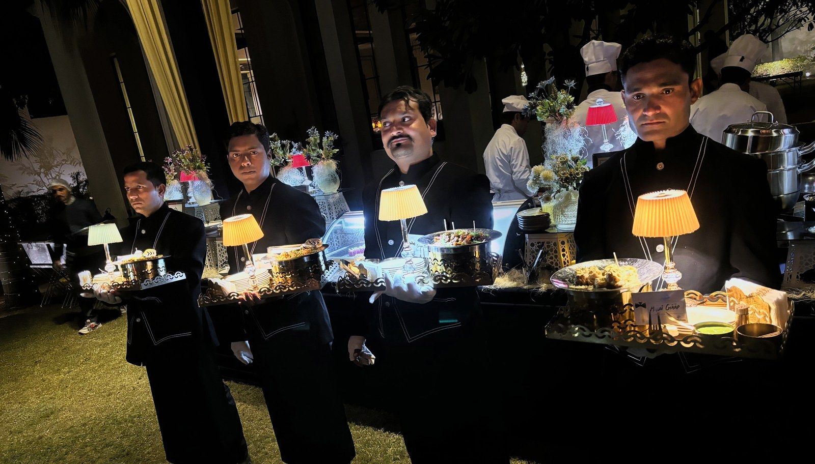 Four servers carrying lamp-lit trays across a lawn